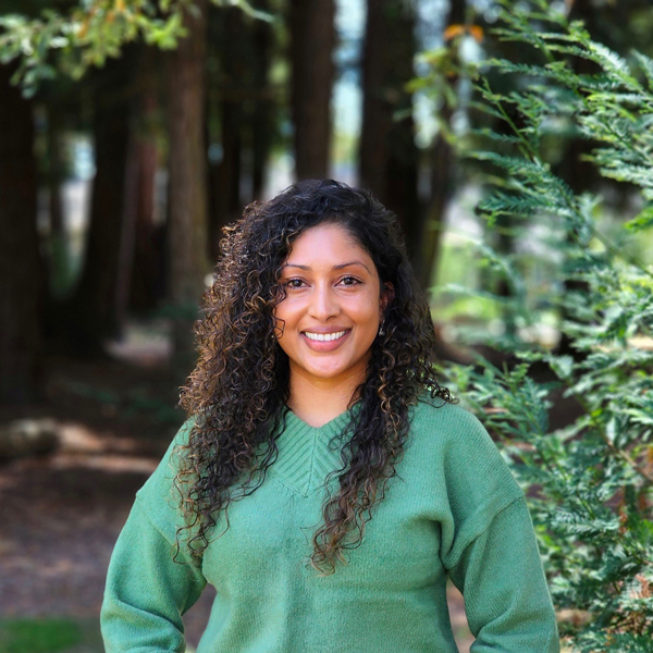 Swasthi Naidu, Mental Health Supervisor at Student Health Services, smiling at the camera while standing outdoors in a forest. She has curly dark hair and is wearing a green sweater.