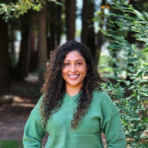 Swasthi Naidu, Mental Health Supervisor at Student Health Services, smiling at the camera while standing outdoors in a forest. She has curly dark hair and is wearing a green sweater.